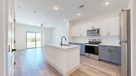 This well-appointed single-floor residence offers a thoughtfully designed open-concept layout that seamlessly connects the kitchen and living area. The kitchen is a standout feature, showcasing a two-tone cabinet design with white upper cabinets and slate-blue lower cabinets, complemented by a large center island with a white quartz countertop and an undermount sink with a matte black faucet. Stainless steel appliances, including a range, microwave, refrigerator, and dishwasher, complete the modern culinary space. Light wood-tone flooring flows throughout the main living areas, enhancing the bright, airy atmosphere created by recessed lighting. The living area opens to a private balcony through a sliding glass door, offering scenic views of the surrounding landscape and natural light. The bedroom features plush carpeting and connects to two closets, including a walk-in closet with wrap-around hanging rods. The bathroom is elegantly finished with a dual-sink vanity, gray cabinetry, matte black hardware, a large mirror, and a glass-enclosed walk-in shower with tile surround. Clean white walls and consistent finishes throughout give the space a cohesive, modern aesthetic.