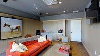 This single-floor space offers a cohesive and thoughtfully designed interior that blends modern style with warm, earthy elements. The main living area immediately draws attention with a vibrant orange tufted sofa that serves as the focal point, complemented by a low-profile coffee table and a pair of cozy accent chairs draped with knit throws. A large area rug anchors the seating arrangement, adding texture and warmth to the hardwood flooring beneath.

The walls are clad in vertical wood paneling with a light, natural finish, lending the space a rustic yet contemporary character. This paneling extends seamlessly along the back wall, framing a built-in kitchenette equipped with a countertop, sink, microwave, and additional small appliances — offering practical convenience within the living environment.

The ceiling is finished in a deep charcoal gray, creating a striking contrast with the lighter wall tones and contributing to the intimate, sophisticated ambiance of the room. Recessed lighting is evenly distributed throughout the ceiling, providing clean, focused illumination, while a decorative ceiling vent adds an architectural detail overhead. A sliding or hinged white door on the side wall provides access to an adjoining area, suggesting additional functionality within the space. Overall, the design is modern, curated, and highly functional.