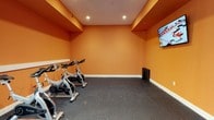 The Cardio Center at 100 Commons Park North in Stamford, Connecticut, is a purpose-built fitness facility designed to support an active and healthy lifestyle. The space is immediately distinguished by its bold, energizing orange walls, which create a motivating atmosphere ideal for cardiovascular workouts. Recessed ceiling lighting provides bright, even illumination throughout the room, ensuring a well-lit environment for exercise at any time of day. The flooring consists of durable rubber gym matting, which offers both safety and comfort underfoot — a practical choice suited to high-intensity fitness activity. A lineup of stationary cycling bikes is positioned along one wall, making the room well-equipped for spin-style workouts. A flat-screen television is mounted on the wall, allowing users to stay entertained or follow along with fitness programming during their sessions. The ceiling features a stepped architectural detail with contrasting trim, adding a subtle design element to the otherwise streamlined interior. The single-floor layout keeps the space focused and accessible, delivering a clean, functional cardio environment within the Commons Park community.