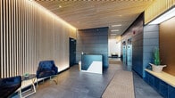 Located at 1367 N Sedgwick St in Chicago's Old Town neighborhood, this single-floor lobby presents a polished, contemporary design that sets a sophisticated tone for the building. The entrance foyer features large-format grey tile flooring, floor-to-ceiling vertical wood slat accent walls with integrated ambient backlighting, and a matching slatted wood ceiling that adds warmth and visual texture throughout the space.

Natural light filters through expansive glass automatic entry doors, illuminating a welcoming seating area furnished with tufted navy velvet chairs and sleek gold-framed accent tables. The transition from the entry vestibule into the main lobby is seamless, guiding visitors toward a prominent reception desk finished in dark blue and white tones, centrally positioned against a backdrop of dark, paneled wall cladding.

The elevator corridor extends along the right side, featuring the same dark metal panel wall treatment and stainless steel elevator doors, complemented by a secured access door for additional building areas. Recessed ceiling lighting and linear LED strips provide consistent, layered illumination throughout. The overall ambiance is modern and refined, combining natural wood tones with dark metallic finishes to create a welcoming yet professional environment.