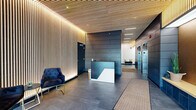 Located at 1367 N Sedgwick St in Chicago's Old Town neighborhood, this single-floor lobby presents a polished, contemporary design that sets a sophisticated tone for the building. The entrance foyer features large-format grey tile flooring, floor-to-ceiling vertical wood slat accent walls with integrated ambient backlighting, and a matching slatted wood ceiling that adds warmth and visual texture throughout the space.

Natural light filters through expansive glass automatic entry doors, illuminating a welcoming seating area furnished with tufted navy velvet chairs and sleek gold-framed accent tables. The transition from the entry vestibule into the main lobby is seamless, guiding visitors toward a prominent reception desk finished in dark blue and white tones, centrally positioned against a backdrop of dark, paneled wall cladding.

The elevator corridor extends along the right side, featuring the same dark metal panel wall treatment and stainless steel elevator doors, complemented by a secured access door for additional building areas. Recessed ceiling lighting and linear LED strips provide consistent, layered illumination throughout. The overall ambiance is modern and refined, combining natural wood tones with dark metallic finishes to create a welcoming yet professional environment.