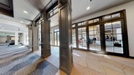 This single-floor amenity facility showcases a refined architectural aesthetic that balances elegance with practicality. The entry lobby sets an impressive tone with expansive natural flagstone flooring laid in irregular patterns, dark-stained wood panel columns, and floor-to-ceiling glass French doors that flood the space with natural light and provide views of the outdoor surroundings. A reception or concierge counter finished in soft gray paneling with a clean white countertop is accented by a geometric hexagonal wallcovering, adding a modern touch to the otherwise classic design palette.

The transition between areas is defined by the consistent use of dark-framed millwork, paneled columns, and transom windows that maintain a cohesive visual language throughout. The fitness center is a standout feature, offering a generously proportioned open floor plan covered in bold hexagonal-patterned carpet tiles in neutral tones of gray and beige. Coffered ceilings with dark wood beams add architectural depth, while recessed lighting and large mirrors enhance the sense of spaciousness. The gym is well-equipped with a variety of strength and cardio machines, supported by columns that delineate workout zones without sacrificing the open feel. Overall, the space presents a polished, community-oriented environment designed for both comfort and active use.