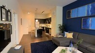 Located at 8110 Six Forks Rd in Raleigh, NC, this single-floor unit presents an open-concept living and kitchen area designed with modern sensibilities. The living space is anchored by a striking navy blue accent wall, which adds depth and character to the room, complemented by warm wood-tone flooring that runs throughout. A sectional sofa, area rug, and curated décor create a comfortable and stylish atmosphere in the living area.

The kitchen is seamlessly integrated into the open floor plan, featuring dark cabinetry that contrasts cleanly with light countertops and stainless steel appliances, including a refrigerator, microwave, and dishwasher. A kitchen island with seating provides a casual dining option and serves as a natural dividing point between the kitchen and living spaces. Pendant lighting above the island adds a refined touch, while track lighting overhead ensures the space is well-illuminated throughout. Multiple doorways visible from the main living area suggest additional rooms beyond, and the overall layout offers a sense of openness and functional flow throughout the space.