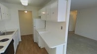 This single-story space features a practical and straightforward layout with a kitchen that serves as a central element of the interior. The kitchen is outfitted with white upper cabinetry offering ample storage, complemented by white lower cabinets and a full-size refrigerator. A notable feature is the extended white countertop that wraps around a partial wall, creating a natural division between the kitchen and the adjacent living area while providing additional workspace or casual dining potential.

The flooring in the kitchen area consists of wood-look planks that add warmth to the space. A textured ceiling is visible throughout, along with flush-mounted ceiling light fixtures that provide functional overhead lighting. The kitchen opens visually into the adjoining rooms, which appear to feature carpeted flooring, contributing to a distinct transition between spaces.

Louvered closet doors are visible in the background, suggesting accessible storage nearby. The overall aesthetic is clean and utilitarian, with a neutral color palette of whites and warm wood tones throughout. The open sightlines between the kitchen and living areas give the space a connected, cohesive feel.