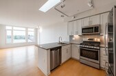 This thoughtfully designed single-floor residence showcases a clean, modern aesthetic throughout its open layout. The kitchen serves as the centerpiece of the living area, highlighted by a striking skylight that floods the space with natural light. White shaker-style cabinetry with sleek hardware lines the walls, complemented by dark gray countertops and a tiled backsplash. A peninsula island with an integrated sink adds both functionality and visual interest, while a full suite of stainless steel appliances — including a gas range, built-in microwave, refrigerator, and dishwasher — elevates the culinary experience. Track lighting runs along the ceiling, providing adjustable illumination throughout. Light-toned hardwood flooring extends seamlessly across the main living areas, contributing to an airy, cohesive feel. A bedroom features large, floor-to-ceiling windows that invite abundant natural light and offer open views of the surrounding neighborhood. The bathroom is finished with dark slate tile flooring, white cabinetry with matching gray countertops, and a narrow vertical window that provides natural light while maintaining privacy. The overall design balances contemporary finishes with a bright, welcoming ambiance.