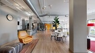 Located at 6250 Griffin Rd in Davie, FL, this single-floor commercial space offers a generously sized, open-plan layout defined by a modern industrial aesthetic. Exposed ductwork runs across the ceiling, complementing the wood-look tile flooring and structural columns that punctuate the interior. Bold accent walls in teal and amber yellow add vibrant visual interest throughout.

The space is organized into several distinct functional zones. A lounge area near the entrance features leather sofas, armchairs, and a round coffee table set atop a red area rug, anchored by large floor-to-ceiling windows that flood the area with natural light and offer outdoor views. Adjacent to this is a collaborative seating zone with high bar-height tables and modern stools, ideal for informal gatherings or focused work.

A well-appointed kitchenette includes a long counter with bar seating, glass-front cabinetry, white subway tile backsplash, stainless steel appliances, and pendant lighting overhead. Additional dining-style tables with contemporary white chairs provide flexible seating options nearby.

Track lighting, pendant fixtures, and exposed mechanical systems overhead contribute to the space's industrial-modern character. Lush indoor plants are strategically placed throughout, enhancing the overall ambiance with a fresh, welcoming atmosphere.