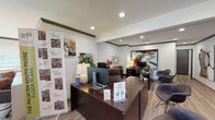 This single-floor leasing office is thoughtfully designed to welcome prospective residents with a polished, professional atmosphere. The main reception area anchors the space, featuring a substantial dark wood front desk equipped with workstations, warm table lighting, and lush potted plants that add a welcoming touch. Bold, contemporary artwork adorns the white walls throughout, complemented by dark crown molding that frames each room with refined detail. Recessed lighting provides consistent, even illumination across the light wood-look flooring that flows seamlessly from room to room. Guest seating with modern chairs is positioned near the reception desk, creating an inviting area for consultations. Leasing information displays and floor plan banners are prominently featured, offering visitors a clear overview of available residence options. A secondary workstation area with a dedicated desk and monitor provides additional staff capacity. Beyond the main office, a bright lounge area serves as a resident amenity space, highlighted by a striking bold orange accent wall, a large sectional sofa, and a refreshment station with a refrigerator and coffee setup. Large windows fitted with vertical blinds allow natural light to filter in throughout the space. Private rooms accessible via gray interior doors offer additional meeting or office space, completing this fully functional and visually engaging leasing office environment.