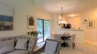 This single-floor residence offers a thoughtfully designed open-concept layout that seamlessly connects the living, dining, and kitchen areas. The living space is warm and inviting, filled with natural light that streams through large sliding glass doors, which provide direct access to a private outdoor patio surrounded by mature, verdant trees — creating a peaceful, nature-immersed backdrop.

The dining area sits adjacent to the living room and is anchored by a dark circular dining table paired with upholstered chairs featuring nailhead trim detailing. A elegant multi-arm chandelier with frosted glass shades hangs overhead, adding a refined touch to the space.

The kitchen is positioned just beyond the dining area and features a raised pass-through counter that maintains visual connectivity with the rest of the open floor plan. White upper cabinetry and black appliances, including a refrigerator and range, give the kitchen a clean, classic contrast. A small shelf along the pass-through provides additional display or prep space.

Throughout the main living areas, light-toned carpet and soft, neutral wall colors create a cohesive and calming ambiance. The overall design balances comfort and functionality, with abundant natural light and greenery views enhancing the sense of openness throughout the home.