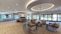 The amenity level at Renaissance Bay Residences, located at 200 La Bonne Vie Drive in Patchogue, New York, is a thoughtfully designed single-floor facility offering residents a refined collection of communal spaces. The centerpiece is a expansive lounge area characterized by a coffered ceiling with integrated ambient lighting, a dramatic floor-to-ceiling marble fireplace surround, and plush seating arrangements set atop patterned carpeting. Large glass doors and windows flood the space with natural light while providing views of the surrounding greenery. An adjoining gathering area features a distinctive circular tray ceiling detail and additional lounge seating, creating an inviting atmosphere for socializing.

The fitness center is well-appointed with rows of treadmills, strength-training cable machines, free weights, and a dedicated area with a heavy punching bag and functional training equipment. Full-length mirrors and ceiling fans contribute to a comfortable workout environment, while large windows offer natural light and outdoor views.

A sleek catering kitchen features white quartz countertops, a multi-basin stainless steel sink, and marble-tiled backsplash accents with linear LED lighting. A separate package room with dark-paneled lockers and light wood-style flooring provides practical storage convenience for residents.