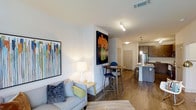 This single-floor space showcases a thoughtfully designed open-concept layout that integrates the living, dining, and kitchen areas into one cohesive, flowing environment. Rich hardwood-style flooring runs throughout, providing warmth and continuity across the space.

The living area features a light-toned sofa accented with colorful throw pillows, complemented by vibrant wall art that adds a pop of personality. A softly glowing table lamp enhances the welcoming ambiance.

The dining area is defined by a round wooden table paired with modern bar-height chairs, positioned beneath bold floral artwork that serves as a striking focal point.

The kitchen sits at the far end of the open plan, anchored by a sleek island with bar stool seating. Dark cabinetry provides contrast against lighter countertops, while stainless steel appliances and pendant lighting lend a contemporary, polished feel.

Recessed and track lighting throughout ensures the space is well-lit, while crisp white walls keep the overall atmosphere bright and airy. The open flow between zones makes this a highly functional and visually cohesive space.