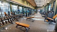 Located at 229 E Chatham in Cary, NC, this single-floor fitness center is a well-appointed exercise facility designed to support a comprehensive workout experience. The main exercise area features a striking architectural aesthetic, with deep olive-green accent walls, a slatted wood ceiling detail, and stylish pendant lighting that lend the space a modern, upscale ambiance. Durable speckled rubber flooring spans the primary workout zone, providing a practical yet polished surface for strength training and cardio activity. Large, floor-to-ceiling windows along the perimeter flood the interior with abundant natural light and offer open views to the surrounding streetscape and outdoor areas. The facility is generously equipped with a full rack of dumbbells, multiple adjustable benches, cable and resistance machines, treadmills, ellipticals, and additional cardio equipment. Ceiling fans and mounted flat-screen televisions are positioned throughout for comfort and entertainment. A separate studio room, finished with light hardwood flooring and soaring ceilings, provides space for functional training and group exercise, complete with a mirrored wall, battle ropes, step platforms, and a large display screen. The overall atmosphere is energetic yet refined, combining thoughtful design with a thorough selection of fitness amenities.