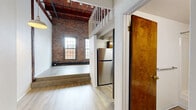 Located at 70 Union Place in Hartford, CT, this loft-style unit offers a compelling blend of industrial heritage and contemporary updates. The main living area immediately captures attention with soaring ceilings featuring rich, dark-stained wooden beams and planking, paired with exposed brick walls that speak to the building's historic origins. Large arched and grid-pane windows line the brick wall, drawing in generous natural light and framing views of the surrounding urban landscape.

Light-toned hardwood-style flooring flows throughout the main level, complementing the rawness of the brick and wood overhead. A white-railed staircase leads to an open mezzanine level above, providing additional functional space overlooking the living area below—a defining architectural feature of the unit's loft character. Exposed piping runs along the ceiling, reinforcing the industrial aesthetic.

The galley-style kitchen is efficiently laid out with warm wood cabinetry lining both sides, dark countertops, stainless steel appliances including a full-size refrigerator and range, and a skylight panel above for added brightness. The bathroom is clean and straightforward, featuring wood-look tile flooring, a full tub/shower combination, and simple, functional fixtures. The unit's overall ambiance is open, airy, and characterful.