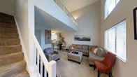 This two-story residence makes a striking impression with its dramatic vaulted ceilings that soar above the main living area, creating an expansive and airy atmosphere throughout the home. The living room serves as the central gathering space, characterized by its generous proportions, plush carpeting, and an abundance of natural light streaming in through large windows. A comfortable sectional sofa and coordinating seating are arranged around a central ottoman, making the space ideal for both relaxing and entertaining. A large landscape artwork adorns the wall, adding a decorative focal point to the room. A staircase with white-painted railings connects the two floors and features an open upper-level landing that overlooks the living area below, enhancing the sense of vertical space and connectivity between floors. Adjacent to the living room, a dining area with a bistro-style table and chairs transitions naturally into the kitchen, which is visible in the background with its clean, light-toned cabinetry and functional layout. The open-concept design of the main floor ensures a smooth flow between living, dining, and kitchen areas, making the space feel cohesive and well-integrated throughout.