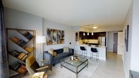 This thoughtfully designed single-floor space showcases a seamless open-concept layout that connects the living area and kitchen into one cohesive, functional environment. The living room is anchored by a plush tufted charcoal sofa, a glass-top coffee table with metallic accents, and a statement abstract canvas that adds bold color to the neutral gray walls. A gold-toned floor lamp and decorative shelving unit contribute warmth and visual interest to the space. Light-toned flooring runs throughout, enhancing the sense of openness and brightness. The kitchen is a standout feature, boasting dark cabinetry, pendant lighting, a stainless steel refrigerator, and a white countertop island with bar seating for two — ideal for casual dining or entertaining. Track lighting overhead ensures the kitchen is well-lit and functional. The overall aesthetic blends contemporary and transitional design elements, with a curated palette of gray, gold, and black that creates a polished, upscale atmosphere. The open flow between spaces makes the layout feel expansive and inviting, well-suited for both everyday living and hosting guests.