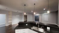 This single-floor residence offers a thoughtfully designed open-concept layout that seamlessly connects the kitchen and living areas, creating a spacious and airy atmosphere. The kitchen serves as the centerpiece of the home, showcasing striking dark granite countertops that contrast beautifully against crisp white cabinetry. A stainless steel sink is elegantly integrated into the countertop, complemented by a modern pull-down faucet. Two globe-style pendant lights hang above the kitchen island, adding a warm, contemporary touch to the space. Recessed lighting throughout further enhances the bright, well-lit ambiance. The open living area extends naturally from the kitchen, bathed in soft light and featuring neutral gray walls that lend a clean, modern aesthetic. A window dressed with flowing white curtains allows natural light to filter in, offering a subtle connection to the outdoors. Hardwood-style flooring runs throughout, tying the spaces together cohesively. An additional linear pendant light fixture is visible in an adjacent dining or flex area, hinting at the versatility of the floor plan. Overall, the space combines modern finishes with a functional, open design that is both inviting and sophisticated.