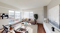 This well-appointed upper-floor residence offers a thoughtfully designed open-concept layout that seamlessly connects the main living areas. The spacious living room is bathed in natural light from large floor-to-ceiling windows, which frame expansive urban views of the surrounding cityscape. Rich hardwood flooring runs throughout the main living space, adding warmth and continuity to the design.

The kitchen is both functional and stylish, featuring light maple cabinetry, stainless steel appliances including a refrigerator, dishwasher, and range, as well as a white countertop island that doubles as a casual dining and prep area. Track lighting overhead provides ample illumination throughout.

The primary bedroom is generously sized with plush carpeting, a large picture window showcasing city views, and a full en-suite bathroom. The en-suite bath includes a dual-sink vanity with maple cabinetry and hardwood floors. A second well-proportioned bedroom also features carpeted floors, a double-door closet, and natural light. The second full bathroom offers a walk-in shower, a single vanity, and hardwood flooring.

Overall, the residence presents a clean, contemporary aesthetic with neutral tones, abundant natural light, and elevated urban views throughout.