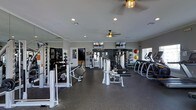 This single-floor fitness center is thoughtfully designed to support a full range of workout routines. The space is outfitted with an impressive array of strength training equipment, including cable machines, weight benches, and a dumbbell rack, alongside cardio machines such as treadmills and ellipticals positioned along the perimeter. Large mirrored walls line multiple sides of the room, creating a sense of expansiveness while allowing exercisers to monitor their form. Overhead, ceiling fans with decorative gold-toned fixtures provide consistent airflow throughout the space, contributing to a comfortable environment during intense workouts. Recessed lighting supplements the natural light that filters in through windows along the walls. Wall-mounted flat-screen televisions are positioned at eye level, offering entertainment options for those using the cardio equipment. The flooring features a durable, dark rubberized surface suited for heavy equipment and high-traffic use. Medicine balls and additional accessories are neatly organized along the walls, ensuring the space remains functional and accessible. The overall ambiance is clean, modern, and well-maintained, making it a practical and inviting facility for fitness enthusiasts.
