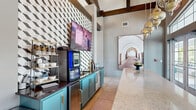 Located in Naples, Florida, this single-floor community space offers a thoughtfully designed interior that blends contemporary style with warm, eclectic touches. The entrance immediately impresses with soaring vaulted ceilings accented by exposed dark wood beams, flooding the space with an airy, open ambiance. Towering floor-to-ceiling windows along the front façade invite abundant natural light, while French doors provide seamless access to the outdoors.

A standout feature is the elongated reception counter finished in polished marble-style stone, anchored by a rich mauve base and illuminated from above by a cascading row of ornate Moroccan-inspired pendant lights. Adjacent to the reception area, a well-appointed refreshment station showcases bold teal cabinetry topped with dark granite countertops, a geometric three-dimensional tile feature wall in black, white, and grey, and a modern self-serve coffee machine complete with a cup and supply organizer.

Throughout the space, curated artwork and vibrant gallery walls add personality, while large-format photography pieces and colorful framed prints create visual interest at every turn. Large format tile flooring unifies the layout and complements the bright, modern palette. The adjacent lounge area, partially visible through a wide corridor, continues the lively and welcoming aesthetic, making this a functional yet inviting community hub.