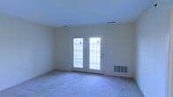 This single-floor space features a straightforward, functional layout centered around a bright main living area. The room is finished with wall-to-wall carpet in a neutral beige tone, providing a soft underfoot feel that contributes to a quiet, comfortable atmosphere. The walls are painted in a light, creamy off-white shade, creating a clean and airy backdrop throughout.

A key architectural feature of this room is the exterior door paired with a sidelight window, both fitted with horizontal blinds that allow for adjustable natural light control. This door-and-window combination serves as the primary source of natural light in the space, brightening the interior while also providing direct exterior access. The door's white frame contrasts cleanly against the surrounding walls.

The ceiling is smooth and standard in height, with flush-mounted HVAC vents positioned for efficient air distribution. A floor-level wall vent further supports climate control. The overall design is simple and practical, with no ornate detailing, making the space highly versatile and easy to configure for a variety of uses. The neutral palette and carpeted flooring lend the room a calm, understated character.
