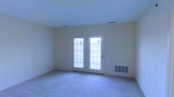 This single-floor space features a straightforward, functional layout centered around a bright main living area. The room is finished with wall-to-wall carpet in a neutral beige tone, providing a soft underfoot feel that contributes to a quiet, comfortable atmosphere. The walls are painted in a light, creamy off-white shade, creating a clean and airy backdrop throughout.

A key architectural feature of this room is the exterior door paired with a sidelight window, both fitted with horizontal blinds that allow for adjustable natural light control. This door-and-window combination serves as the primary source of natural light in the space, brightening the interior while also providing direct exterior access. The door's white frame contrasts cleanly against the surrounding walls.

The ceiling is smooth and standard in height, with flush-mounted HVAC vents positioned for efficient air distribution. A floor-level wall vent further supports climate control. The overall design is simple and practical, with no ornate detailing, making the space highly versatile and easy to configure for a variety of uses. The neutral palette and carpeted flooring lend the room a calm, understated character.