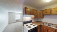 This single-floor unit offers a practical and straightforward layout designed for comfortable everyday living. The kitchen is efficiently arranged in an L-shaped configuration, featuring warm oak cabinetry with decorative hardware, laminate countertops, a double-basin stainless steel sink, a coil-top electric range, and a white refrigerator. A ventilation hood is mounted above the range, and durable wood-look vinyl flooring runs throughout the kitchen and entry areas.

The living space transitions from the kitchen and is finished with neutral wall-to-wall carpet, providing a softer feel underfoot. A ceiling fan with an integrated light fixture is present in the entryway area, which also features baseboard heating and a straightforward front door entry. The open layout allows for flexible furniture arrangements.

The bedrooms are carpeted and finished with neutral tones, each featuring individual closets with hanging rods. The primary bedroom includes two closets, a window for natural light, and a wall-mounted air conditioning unit alongside baseboard heating. A second bedroom offers a single closet and additional carpeted space.

The bathroom is clean and functional, equipped with a full-sized soaking tub and shower combination with white tile surround, complemented by wood-look flooring. Throughout, textured ceilings add subtle visual detail to the space.