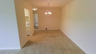 This single-floor residential space offers a straightforward and functional layout with a warm, neutral aesthetic throughout. The main living and dining area is generously sized, featuring wall-to-wall beige carpeting that lends a soft, cozy feel underfoot. A classic chandelier with bell-shaped shades serves as a central focal point in the dining area, adding a touch of traditional elegance to the space. The walls are finished in a soft, warm tone that complements the light carpeting and contributes to an overall bright and welcoming ambiance. A baseboard heating unit lines one wall, providing efficient climate control throughout the colder months. An open doorway connects the living area to the kitchen, which is equipped with white cabinetry and standard appliances, maintaining a clean and practical design. A hallway visible beyond the kitchen suggests additional rooms, offering a logical and easy-to-navigate floor plan. The space is currently vacant, making it easy to envision a variety of furniture arrangements and personal touches. Overall, the unit presents a clean, move-in ready environment with a simple yet functional design suitable for a range of lifestyles.