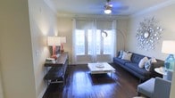 This single-floor space offers a thoughtfully designed interior centered around a welcoming living room. Rich, dark hardwood floors run throughout, providing a warm and polished foundation that complements the neutral-toned walls. Large, multi-pane windows dressed with sheer white curtains allow an abundance of natural light to pour in, creating an airy and inviting atmosphere. Crown molding along the ceiling adds a refined architectural touch, while a ceiling fan with an integrated light fixture provides both style and function. The living area is well-proportioned, offering ample room for comfortable furnishings. A sleek, modern sofa anchors the seating arrangement alongside a minimalist white coffee table, while a rustic console table along the wall adds contrast and character. An arc floor lamp introduces a contemporary design element, complementing the overall aesthetic. The space strikes a balance between classic architectural details and modern interior styling, making it both functional and visually appealing. The generous window presence ensures the room feels open and connected to the outdoors, enhancing the overall sense of brightness and space throughout the home.