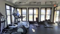 This fitness center is a dedicated, single-floor space designed to support a comprehensive workout experience. The room is lined with large, dark-framed windows along one wall, allowing abundant natural light to pour in and offering views of the surrounding greenery — creating an open, energizing atmosphere for exercise.

The flooring is finished with durable rubber gym matting, providing both safety and functionality underfoot. Overhead, a track lighting system ensures the space remains well-lit throughout the day and into the evening.

The cardio section is well-appointed, featuring multiple treadmills — including a Life Fitness model — alongside an elliptical cross-trainer, giving users a variety of low- and high-impact aerobic options. The strength training area includes a weight bench and a cable/resistance machine, catering to users focused on muscle conditioning and targeted training.

The layout is thoughtfully organized, with equipment arranged to allow comfortable movement between stations. The combination of natural light, outdoor views, and a well-rounded selection of fitness equipment makes this a functional and motivating environment for residents or members looking to maintain an active lifestyle.