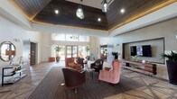 Located at 12325 Shadow Creek Pkwy in Pearland, Texas, this single-floor clubhouse facility is designed with a refined, contemporary aesthetic that balances warmth and sophistication. The grand entry area immediately impresses with its dramatic vaulted ceiling clad in rich dark wood paneling, complemented by statement pendant lighting and recessed fixtures that cast a welcoming glow throughout the space.

One of the most striking architectural features is the expansive geometric hardwood floor inlay, forming an intricate starburst pattern that anchors the central gathering area. This main lounge is furnished with a curated mix of upholstered seating — including plush pink wingback chairs, leather armchairs, and patterned dining-height chairs — arranged to encourage both casual conversation and formal seating.

Floor-to-ceiling windows and glass doors along the perimeter flood the interior with abundant natural light and offer views of the surrounding outdoor landscape. A sleek, modern fireplace framed in smooth concrete serves as a focal point in one lounge area, flanked by wood-framed chairs and decorated with sculptural wall art above the mantle.

Sliding barn-style doors in warm wood tones provide passage to additional connected rooms, including what appears to be a business center and a secondary lounge or multipurpose room. A wall-mounted flat-screen television and built-in stone-accented media console add functional amenities to the communal areas. The overall ambiance is polished yet inviting, reflecting a well-appointed community facility.