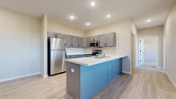 This well-appointed single-floor residence offers a thoughtfully designed open-concept layout that seamlessly connects the kitchen and living areas. The kitchen is a standout feature, showcasing a bold blue peninsula with a white countertop, grey shaker-style cabinetry with black hardware, and a full suite of stainless steel appliances including a refrigerator, range with over-the-range microwave, and dishwasher. A white tile backsplash and recessed lighting complete the space with a clean, modern aesthetic. Light wood-look flooring flows throughout the main living areas, complementing the neutral wall tones and creating a bright, cohesive atmosphere. The living area is generously proportioned, featuring a ceiling fan and windows that invite natural light while providing views of the surrounding outdoors. The bedroom is carpeted for comfort, equipped with a wall-mounted HVAC unit and a large window. The bathroom features a wide grey vanity with ample storage, white countertop, and a large mirror. A carpeted room with an electrical panel offers flexible utility use. The attached garage is spacious, drywalled, and equipped with an automatic door opener, providing ample room for vehicles and storage. Throughout, the space reflects a modern, move-in-ready design with consistent finishes.