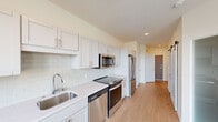 This single-floor unit showcases a clean, contemporary design with thoughtful finishes throughout. The kitchen is a standout feature, outfitted with white shaker-style cabinetry, light quartz countertops, and a subtle subway tile backsplash. A full suite of stainless steel appliances — including a refrigerator, electric range, built-in microwave, and dishwasher — provides a polished, functional workspace. Light hardwood-style flooring extends through the kitchen and into the open living area, which benefits from generous natural light through large windows fitted with horizontal blinds. A sliding barn door adds a stylish architectural accent along the hallway. The bedroom features plush carpeting underfoot, a tall frosted window that allows diffused natural light, and a wide double sliding-door closet offering substantial storage. The bathroom is well-appointed with a white vanity, undermount sink, quartz countertop, large wall mirror with overhead lighting, and a glass-enclosed walk-in shower. Recessed lighting and neutral tones throughout contribute to a bright, cohesive ambiance across the entire space.