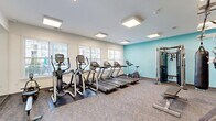 Located at 14300 Sapphire Park Ln in Midlothian, Virginia, this single-floor fitness center offers a well-appointed workout facility designed to accommodate a wide range of exercise routines. The space is divided into distinct functional zones, each equipped to support different training styles.

The main workout area features a row of treadmills, elliptical trainers, and an upright cycle, all positioned along a bank of windows that allow ample natural light to fill the room. A signature teal accent wall adds a vibrant pop of color to the otherwise clean, neutral palette. Overhead, flush-mounted panel lighting ensures consistent illumination throughout.

The strength and functional training zone is anchored by a RAX multi-functional cable and suspension training system, complemented by a full rack of kettlebells and dumbbells, a punching bag, an adjustable workout bench, and a wall-mounted flat-screen television. Mirror panels line the walls, enhancing the sense of space and supporting form monitoring during exercise. A dedicated stretching and agility area with floor markings is also present.

A separate spin studio rounds out the facility, housing multiple Matrix cycling bikes arranged before a large wall mirror and mounted display screen. This room features a bold branded mural in purple and blue tones, a window with outdoor views, and smooth light-toned flooring, creating an energetic yet focused atmosphere for cycling workouts.