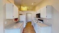 Located at 2415 N Cherry St in Spokane Valley, WA, this single-floor unit offers a well-organized, open-concept layout designed for comfortable everyday living. The galley-style kitchen is efficiently arranged with ample white cabinetry, light granite-style countertops, wood-look flooring, a double-basin sink, and a full suite of appliances including a refrigerator, electric range, dishwasher, and over-the-range microwave. Vaulted ceiling accents above the kitchen add visual interest and an airy feel to the space. A pass-through opening connects the kitchen to the dining area, which features a pendant light fixture and a window that brings in natural light. The living room is generously proportioned with plush carpeting, vertical blinds covering a sliding glass door, and a wall-mounted air conditioning unit. Bedrooms are carpeted and feature neutral wall tones alongside sliding wood-panel closet doors offering practical storage. The bathroom includes wood-look flooring, a full soaking tub with shower, and clean, neutral finishes throughout. The unit's entry is accessed through an arched doorway, adding a subtle architectural touch to the overall design.