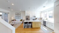 This well-designed two-floor home offers a thoughtful layout with modern finishes and a bright, airy atmosphere throughout. The open-concept main living area seamlessly connects the kitchen and living room, anchored by light hardwood flooring that flows throughout the space. The kitchen is a standout feature, boasting crisp white shaker cabinetry, a classic subway tile backsplash, stainless steel appliances, and a central island with a contrasting soft yellow base and white quartz countertop — complete with bar seating and pendant lighting overhead. The adjacent living area provides direct outdoor access through a sliding glass door leading to a private balcony or patio. A dedicated dining area accommodates a full dining set beneath a striking pendant light, complemented by bold artwork. The bedrooms are generously proportioned, bathed in natural light from large windows, and feature ceiling fans and hardwood floors. The primary bathroom is a luxurious retreat with a double-sink vanity, marble-veined accent wall, walk-in tile shower, hexagonal tile flooring, and an adjoining walk-in closet. A secondary bathroom features charming blue floral wallpaper, hexagonal tile floors, a soaking tub, and classic white cabinetry. An additional flexible room offers versatile living space, ideal as a lounge, playroom, or home office.