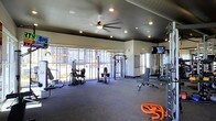 This single-level fitness center offers a comprehensive workout environment designed to support a range of exercise routines. The space features dark-painted ceilings with recessed lighting and a ceiling fan that contribute to an energized yet comfortable atmosphere. Large floor-to-ceiling windows along one wall flood the interior with natural light while providing views of the surrounding exterior, creating an open and motivating environment. The rubber flooring throughout the space is ideal for heavy equipment use and provides cushioning and durability underfoot. The facility is outfitted with multiple cable and weight machines, a dedicated free weights area, and adjustable benches, catering to both strength training and functional fitness. Battle ropes are also available on the floor, adding variety to workout options. Wall-mounted televisions are positioned for easy viewing during exercise sessions. A large mirror along one wall enhances the sense of space and allows users to monitor their form. Overall, the fitness center balances functionality with a well-lit, spacious design that supports an effective and focused workout experience.