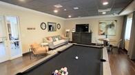 This recreational lounge is thoughtfully designed to serve as a versatile social and entertainment hub. At the heart of the space sits a full-size pool table with a sleek black felt surface, inviting guests to enjoy a friendly game at any time. Recessed lighting set into a drop-panel ceiling illuminates the room evenly, creating a warm and inviting atmosphere throughout.

Adjacent to the pool table, a comfortable seating area features a neutral-toned sofa accented with colorful throw pillows, complemented by a pair of modern accent chairs. A striped area rug anchors the lounge seating, adding visual interest to the hardwood-style flooring beneath. Decorative round mirrors mounted on the warm beige walls lend an elegant, finished touch to the space.

Opposite the seating area, a large flat-screen television is positioned atop a dark wood media console, making the room equally suited for watching sports or movies. Natural light filters in from an adjacent area, adding brightness to the overall environment. A small high-top table and chairs are tucked nearby, offering an additional spot for conversation or casual dining. Together, these elements create a well-rounded, amenity-rich space designed for relaxation and community engagement.