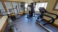 This single-floor fitness space is designed to support a well-rounded exercise routine. The room is outfitted with a variety of equipment, including a treadmill, elliptical machine, stationary bike, and a multi-function cable/weight station, providing options for both cardiovascular training and strength work. The flooring consists of durable speckled rubber matting, a practical choice that offers cushioning and slip resistance during workouts. Multiple large windows with wood-trimmed frames line the walls, allowing natural light to fill the space and offering views to the exterior, contributing to an open and energizing atmosphere. The walls are painted in a soft yellow tone, adding warmth to the utilitarian setting. A decorative potted palm adds a touch of greenery, softening the functional character of the room. The layout is thoughtfully arranged to allow comfortable movement between equipment without crowding. Overall, the space presents a clean, functional environment suited for regular fitness use.