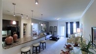 This single-floor space showcases a thoughtfully designed open-concept layout that integrates the kitchen, dining, and living areas into one cohesive and functional environment. The kitchen features a granite-topped breakfast bar with stylish pendant lighting overhead, offering both a practical workspace and a casual seating area with bar stools. A built-in shelving unit adjacent to the kitchen provides additional storage and display space, adding both utility and character to the area. The dining space flows naturally from the kitchen, complete with a glass-top table and upholstered seating. The living area is generously proportioned, finished with plush carpeting and anchored by a tufted ottoman coffee table surrounded by a comfortable sofa and accent chairs in coordinating patterns. Decorative wall art and tasteful accessories lend a polished, curated feel to the space. Crown molding details add an elegant architectural touch throughout. A set of full-length glass doors floods the living area with natural light while offering access to the outdoors, complemented by floor-length curtains that frame the view. A glimpse into the bedroom reveals a well-lit, comfortable private retreat. The overall ambiance is warm, modern, and inviting.