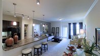This single-floor space showcases a thoughtfully designed open-concept layout that integrates the kitchen, dining, and living areas into one cohesive and functional environment. The kitchen features a granite-topped breakfast bar with stylish pendant lighting overhead, offering both a practical workspace and a casual seating area with bar stools. A built-in shelving unit adjacent to the kitchen provides additional storage and display space, adding both utility and character to the area. The dining space flows naturally from the kitchen, complete with a glass-top table and upholstered seating. The living area is generously proportioned, finished with plush carpeting and anchored by a tufted ottoman coffee table surrounded by a comfortable sofa and accent chairs in coordinating patterns. Decorative wall art and tasteful accessories lend a polished, curated feel to the space. Crown molding details add an elegant architectural touch throughout. A set of full-length glass doors floods the living area with natural light while offering access to the outdoors, complemented by floor-length curtains that frame the view. A glimpse into the bedroom reveals a well-lit, comfortable private retreat. The overall ambiance is warm, modern, and inviting.