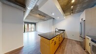 Located at 1025 N Fillmore Street in Arlington, Virginia, this single-floor residence showcases a compelling industrial-modern aesthetic that balances raw architectural elements with refined finishes. The open-concept living and kitchen area is immediately striking, with exposed concrete ceilings, visible ductwork, and track lighting lending an urban loft character to the space. Wide-plank wood-style flooring flows throughout, tying each area together cohesively.

The kitchen is a standout feature, centered around an expansive peninsula topped with dark granite countertops and outfitted with warm natural wood cabinetry accented by matte black hardware. Stainless steel appliances, including a refrigerator, range, and microwave, provide a sleek, functional workspace, while a tile backsplash adds subtle texture. The peninsula opens directly to the generous living area, which benefits from floor-to-ceiling windows that draw in natural light and offer views of the surrounding neighborhood.

The bedroom is a calm, well-proportioned retreat with the same exposed concrete ceiling detail and direct access to natural light via a large window. Two bathrooms serve the unit: one features marble-surround tub and shower with wood-toned cabinetry and granite vanity, while the primary bath offers a marble-clad soaking tub, dual-sink granite vanity with white cabinetry, and recessed lighting, creating a spa-inspired atmosphere.