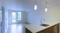 This single-floor space showcases a thoughtfully designed open-concept layout that seamlessly connects the kitchen and living areas. The kitchen features a generous island with a clean white quartz countertop and an undermount stainless steel sink equipped with a sleek arc faucet. Warm wood cabinetry on the island provides a striking contrast to the bright, white surfaces. Two cylindrical pendant lights hang above the island, offering both task lighting and a contemporary design accent.

The living area extends naturally from the kitchen, finished with light-toned hardwood-style flooring that runs throughout, contributing to a cohesive and airy feel. Crisp white walls and a smooth white ceiling enhance the sense of openness and allow natural light to circulate freely. A set of glass-paneled doors at the far end of the living space floods the interior with natural light and provides direct access to an outdoor area, where greenery is visible beyond, adding a refreshing connection to the exterior environment. The overall ambiance is clean, modern, and bright.