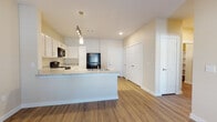 This single-floor residential unit offers a well-designed, functional layout that balances open living spaces with private accommodations. The kitchen serves as a central hub, featuring white shaker-style cabinetry with brushed nickel hardware, light granite-look countertops, and a full suite of black appliances including a refrigerator, electric range, over-the-range microwave, and dishwasher. A peninsula with an integrated sink provides additional prep space and casual seating potential. Pendant and track lighting illuminate the kitchen, which flows seamlessly into the main living area. The living room is bright and open, enhanced by warm wood-look vinyl plank flooring, a ceiling fan with light fixture, and a door providing access to an outdoor area and natural light through blinds-covered windows. The bedrooms are carpeted for comfort and warmth, each featuring windows with blinds and six-panel closet doors offering generous storage. The full bathroom is finished with wood-look vinyl flooring, a vanity with stone-top counters, and a tub-shower combination with tile surround. Neutral warm-beige tones on the walls throughout the unit create a cohesive, clean aesthetic. A walk-in closet or additional storage area is also accessible from the hallway, adding to the unit's overall practicality.