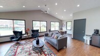 This single-story space is defined by its vaulted ceilings, which lend an airy, expansive feel to the open-concept layout. Recessed lighting and a ceiling fan are positioned throughout, complementing the abundant natural light that pours in through multiple large, black-framed windows offering views of the surrounding greenery outdoors.

The main living area features warm hardwood flooring and is anchored by a gray sofa, a round wooden coffee table, and a vibrant area rug with floral detailing. A pair of navy blue egg-style accent chairs create a dedicated seating nook near the windows, ideal for conversation or relaxation. A large flat-screen television and media console occupy one wall, completing the lounge setup.

Adjacent to the living area, a casual dining nook with a round table and chairs provides an intimate space for meals, while a breakfast bar with barstool seating connects seamlessly to the kitchen. The kitchen is equipped with oak cabinetry, dark granite countertops, stainless steel appliances including a range and microwave, and a stainless sink — offering a practical and well-appointed workspace. The overall ambiance is modern yet comfortable, with neutral tones throughout.