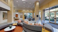 This single-floor amenity space is designed with a modern, open-concept layout that seamlessly connects multiple functional zones beneath soaring ceilings. The main lounge area anchors the space with a large curved sectional sofa in neutral gray, accented by vibrant orange-and-white patterned pillows and set atop a bold orange area rug, creating a warm and inviting focal point. Adjacent to this, a dedicated seating area features upholstered armchairs in complementary orange and gray tones arranged around sculptural marble-topped coffee tables, all bathed in natural light streaming through expansive floor-to-ceiling windows that frame views of lush outdoor courtyard landscaping. A games area equipped with a pool table adds a recreational dimension to the space. High-top bistro tables and bar-height seating provide casual gathering options near the windows. A well-appointed wet bar features warm honey-toned cabinetry, gray tile backsplash with orange accent tiles, integrated appliances, and a mounted flat-screen television. Throughout, warm golden wall paneling, recessed lighting, large-format tile flooring, and carefully curated artwork contribute to a polished, contemporary ambiance ideal for communal use.