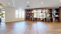 Located at 5121 E La Puente Ave in Phoenix, AZ, this thoughtfully designed single-floor community space offers a generous and versatile living area that seamlessly blends modern aesthetics with warm, eclectic character. The entrance lobby makes an immediate impression with bold geometric patterned tilework, a decorative reception counter with gold mosaic detailing, a striking blue accent piece, and a slatted wood ceiling that adds warmth and texture overhead. Large glass sliding doors and floor-to-ceiling windows flood the interior with abundant natural light while providing views of a furnished outdoor patio, perfect for Arizona's sunny climate. The main living area features rich herringbone wood-look flooring that flows throughout, complemented by an eye-catching carved wood feature wall that anchors the dining and kitchenette zone. Distinctive rattan pendant lights hang above a central bar-height island, while stainless steel appliances and terrazzo-style tile flooring define the kitchen area. A comfortable lounge zone showcases a tufted leather sofa, accent chairs, and curated décor. A study nook features a built-in walnut shelving unit with integrated desk space and multiple windows overlooking the outdoors. Recessed lighting throughout ensures a bright, welcoming ambiance across every zone of this well-appointed space.