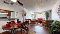 This single-story residence offers a well-organized layout that connects its principal living spaces in a logical, comfortable flow. The living room serves as the heart of the home, featuring a large window dressed with layered gold valance curtains that invite natural light while maintaining privacy. A textured ceiling and ceiling fan add functional character, and the room comfortably accommodates a full seating arrangement anchored by a patterned area rug over wood-look flooring.

Adjacent to the living room, the dining area benefits from a pass-through opening that connects directly to the kitchen, enhancing the open feel of the shared space. The dining area features the same wood-look flooring and provides ample room for a round dining table set. The galley-style kitchen is equipped with rich medium-toned wood cabinetry, dark laminate countertops, a double basin sink, and a full-size refrigerator, offering practical storage and workspace.

The bedroom is a well-proportioned retreat with carpeted flooring, a window with decorative curtains allowing natural light, and a textured ceiling consistent with the rest of the home. The bathroom includes a single-sink vanity with wood cabinetry matching the kitchen, granite-style countertops, a tub-shower combination, and wood-look flooring, completing this cohesive and functional residence.