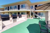 This two-story property is centered around a well-appointed courtyard that serves as its defining feature, offering residents multiple distinct outdoor amenity zones. The courtyard is divided into two functional areas: a garden section and an open activity area.

The garden zone showcases an ornate tiered stone fountain as its centerpiece, surrounded by carefully maintained landscaping including abundant white rose bushes, yellow flowering shrubs, and various potted plants. Wrought-iron tables and chairs are positioned throughout, providing intimate seating areas within the garden setting.

The second courtyard zone features a large artificial turf surface, creating a clean, low-maintenance green space. A built-in outdoor kitchen runs along one side, equipped with multiple BBQ grills set into a tiled masonry counter with storage below — ideal for communal gatherings. Additional wrought-iron patio furniture is scattered across this area.

A central staircase with metal railings provides access to the second-floor balcony walkway, which wraps around the courtyard and leads to individual unit entrances. The building exterior features a warm yellow stucco finish with green accent trim. The entire courtyard receives generous natural light under open skies, enhancing the inviting, resort-like atmosphere.