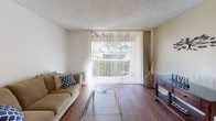 Located at 15513 Williams St in Tustin, California, this single-floor unit offers a well-organized and functional layout designed for comfortable everyday living. The open-concept main area seamlessly connects the living room, dining area, and kitchen, all unified by warm hardwood flooring that runs throughout the common spaces. The living room is bright and inviting, anchored by a large sliding glass door fitted with vertical blinds that allow ample natural light and provide views of the surrounding greenery outside. The kitchen features white cabinetry, a white refrigerator, an electric range, and light countertops, creating a clean and practical cooking space. A peninsula-style counter separates the kitchen from the rest of the open living area. Adjacent to the kitchen, the dining area accommodates a round glass-top table with a ceiling-mounted light fixture and decorative wall art, adding a touch of character to the space. The bedroom is generously sized with plush carpeting, a ceiling fan with integrated lighting, and a window that draws in natural light. A hallway connects the bedroom to additional spaces, including a bathroom visible with light countertops and a mirror. Textured ceilings are consistent throughout the unit, adding a classic residential feel to this well-maintained Tustin home.
