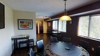 This single-floor space is designed with entertainment and social gathering in mind. The main area centers around a large round poker table with built-in cup holders and plush black leather chairs, making it an ideal setting for game nights. A bold pendant light with a warm amber globe hangs overhead, casting a welcoming glow across the room. The interior features a rich, dark color palette — deep espresso walls contrast with lighter trim and ceilings, complemented by dark hardwood-style flooring throughout. A secondary dining or activity table with bar-height seating is positioned near windows dressed with horizontal blinds, allowing for natural light while maintaining privacy. The textured ceiling adds a classic finish to the space. Colorful abstract artwork adorns the walls, adding personality and visual interest. Toward the back, a hallway leads to additional areas of the space, flanked by dark cabinetry or closet storage. A track lighting fixture provides adjustable illumination along the corridor. The overall ambiance is relaxed yet purposeful — a well-appointed space built for socializing, leisure, and comfortable everyday living.