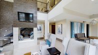 Located at 1701 Atria Cir in Raleigh, NC, this contemporary residence makes an immediate impression with its dramatic double-height living area, where soaring ceilings draw the eye upward to an open mezzanine level with sleek metal railing — a striking architectural focal point. The living space is anchored by a floor-to-ceiling stacked stone accent wall housing a wall-mounted television above a fireplace, lending a bold, textural contrast to the otherwise crisp, white interior. Light-toned flooring and neutral walls create a bright, airy atmosphere throughout, while curated contemporary artwork in cool blue and white tones adds a refined decorative touch. Plush light gray seating arranged around a low-profile coffee table encourages comfortable, casual living. Recessed lighting ensures consistent illumination across the open-plan layout. A deep-set nook within the stone wall offers a cozy, intimate alcove perfect for reading or quiet retreat. Hallways branch off toward additional rooms, providing a functional flow between spaces. The overall design aesthetic is modern and sophisticated, balancing clean architectural lines with warm textural elements to create a polished yet inviting living environment.